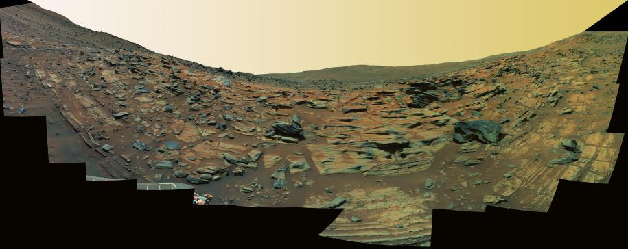 This image shows finely layered rocks interspersed with sand sloping downward and inward toward the center of the panorama from either side. Here and there on the outcrop, a chunk of rock has become displaced and lies at an angle on the surface