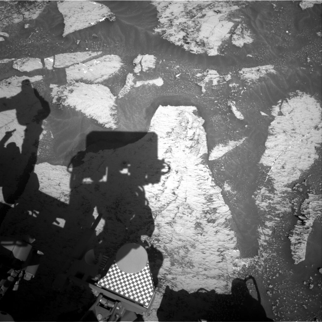 This black and white image shows the Curiosity rover's shadow over a drill hole  on the rocky, sandy surface of Mars.
