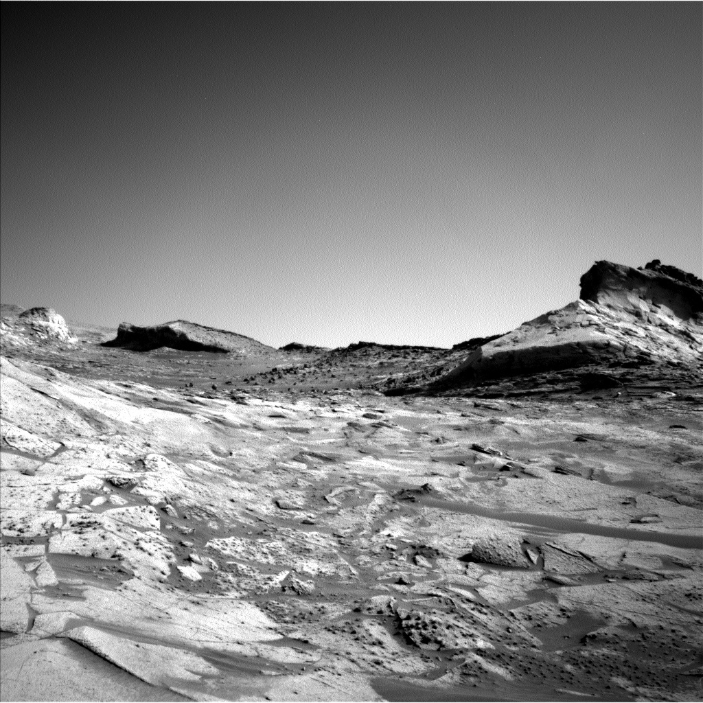 This is a black and white image taken by Left Navigation Camera onboard NASA's Mars rover Curiosity. There are hills in the background and a rocky, sandy surface of Mars is shown in the front of the image. The sky is clear.