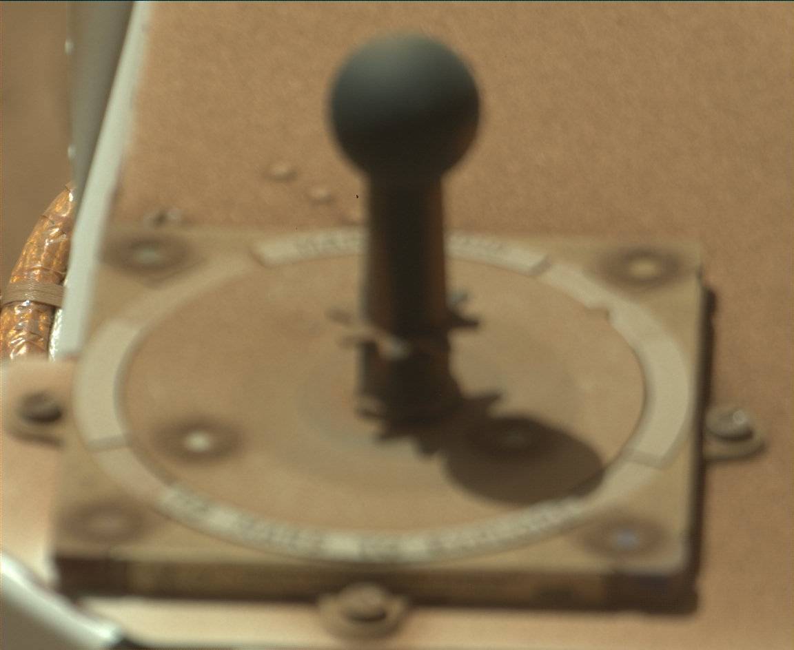 Soft-focus image of the Curiosity rover’s Mastcam calibration target, covered with a thin layer of light brown dust. The target is a small, sundial-shaped installation on top of the rover.
