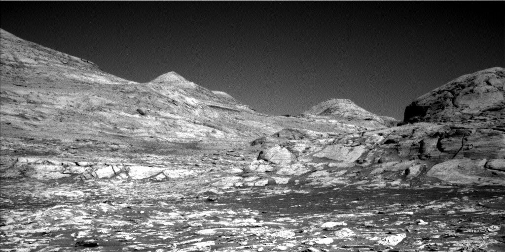 This is a black and white image taken by the Left Navigation Camera of the Rafael Navarro Mountain. The surface of these low hills is very rocky and has cracks and there is a clear sky in the horizon of the image.