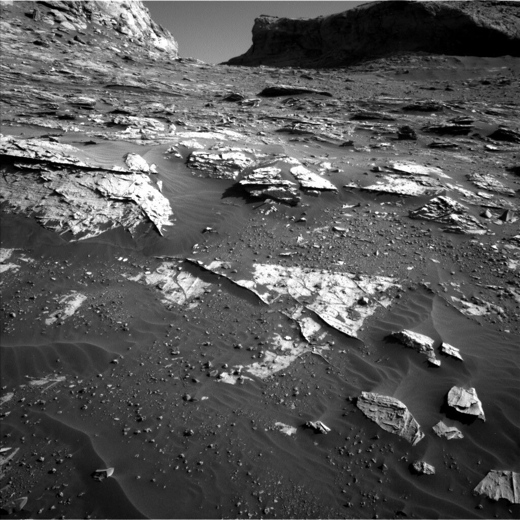 This is a black and white image of the sandy, rocky surface of Mars. There are low hills in the background and small rocks are scattered on the smooth surface.