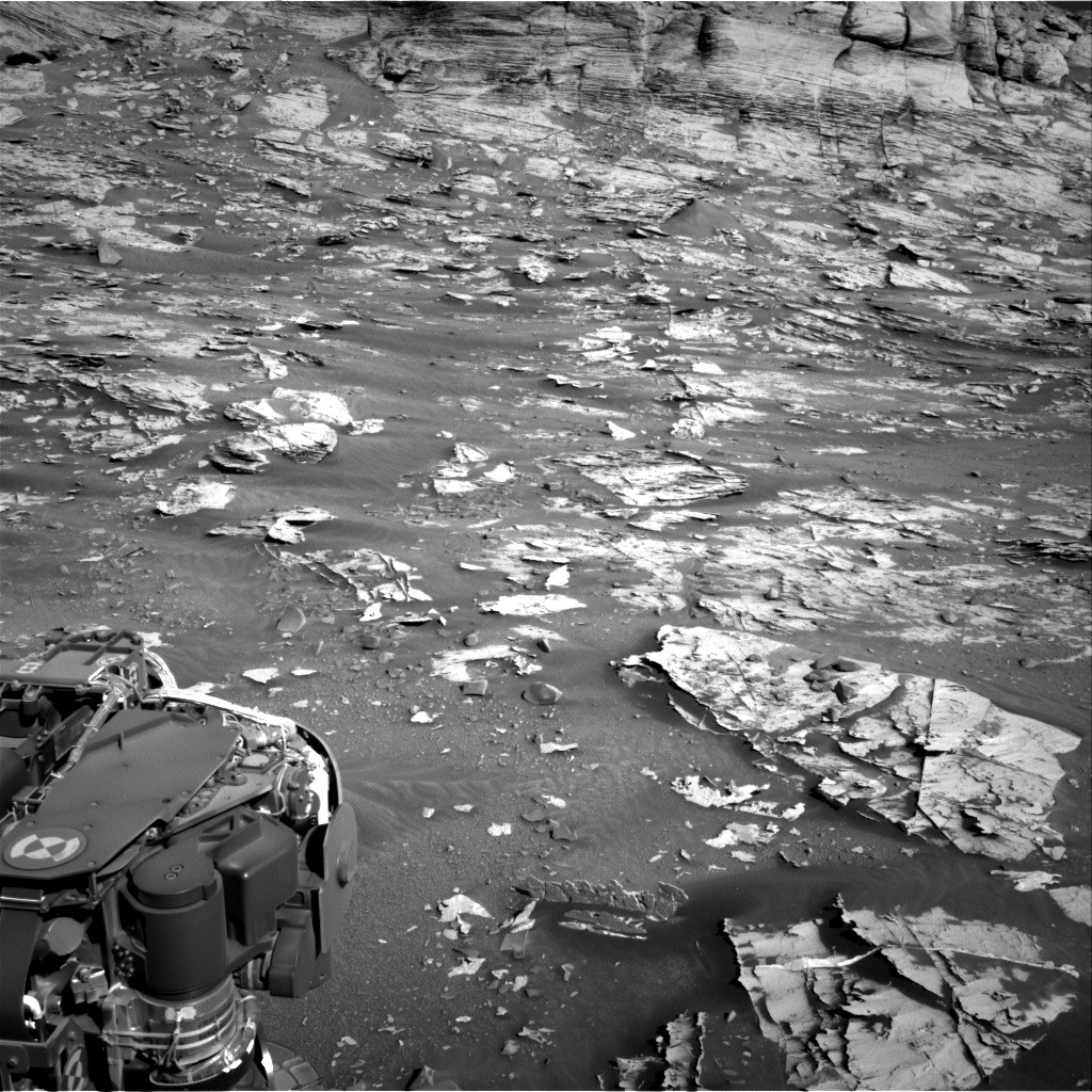 This is a black and white image of the sandy, rocky surface of Mars. There are many flat, low-lying rocks on the surface, and part of the Curiosity rover's arm is visible at lower left.