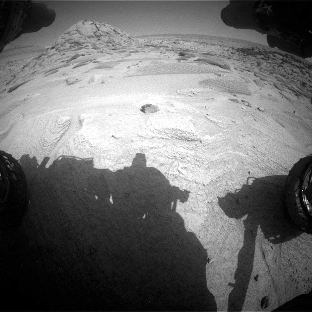 This image shows the shadow of the Curiosity rover above the Mars surface with rocks and a hill in the distance, including float block target "Rio Urubu."