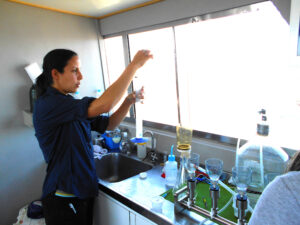 A woman stands to the left of the image, facing the right side, inside a small room and next to a countertop and sink. On the counter are several containers, bottles, and tubing. The woman, wearing a dark blue shirt and black pants, is holding a graduated cylinder out in front of her. There is a window on the back wall in the image, which is casting a bright glow into the room.