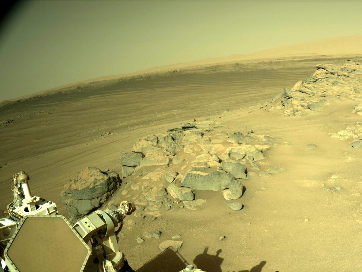 This is an image of the Mars landscape taken by the Perseverance rover. A clear sky, some hills and sandy ground are present int he horizon. Up close there are many large boulders and a part of Perseverance’s AutoNav camera and its shadow are present.