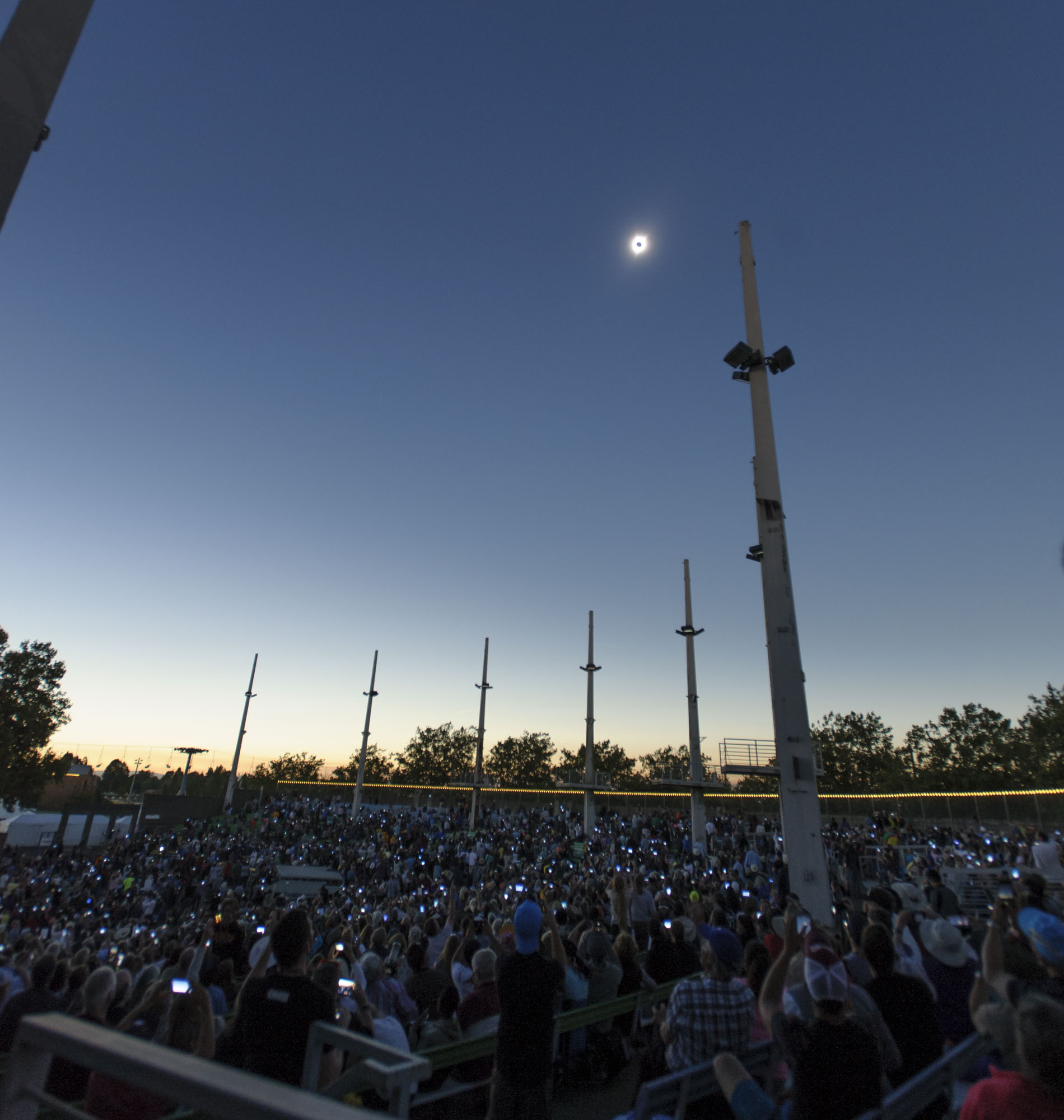 A large crowd of people stand under a totally eclipse Sun. The bright light of their cell phones is seen as they hold them up.