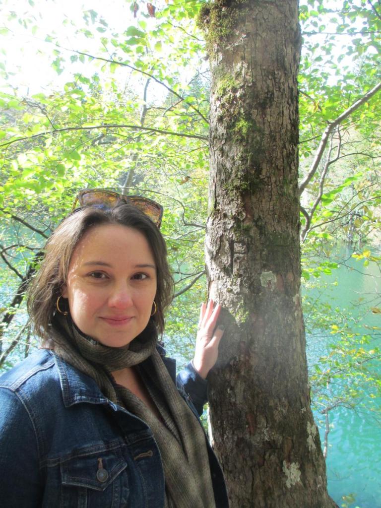 Photo of a young woman with sunglasses resting on top of her head as she stands outside with her hand on a tree trunk.