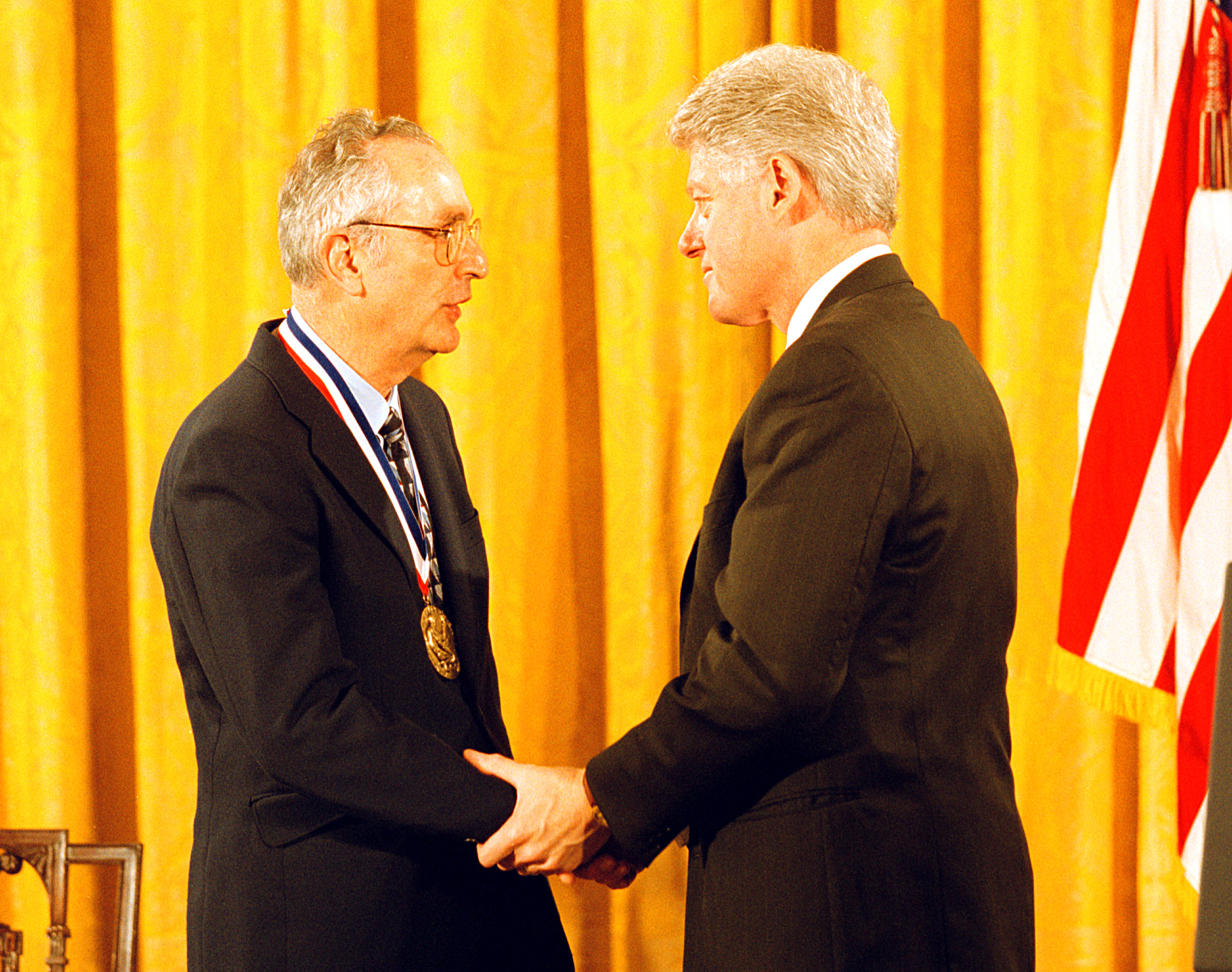 Astronomer John Bahcall (left) receives receives the Medal of Science from President Bill Clinton (right). The two are shaking hands.