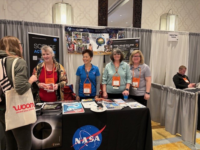 NASA Science Activation team members stand behind a table with assorted handouts and in front of banners.