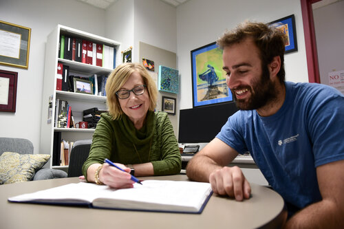Dr. Dorothy Shippen and Dr. Borja Barbero are seated at a round table. They are both looking at a notebook that Dr. Shippen is writing in. Dr. Shippen is donned in a green long-sleeve shirt. She is wearing glasses. Dr. Barbero is wearing a navy-blue t-shirt. A white bookshelf is in the background. Several pieces of artwork and plaques hang on the walls behind them.
