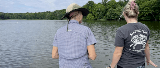 Two women in short sleeved blue shirts stand looking at the water, their backs to the photographer. In front of them is a water, its surface lightly ruffled by wind, and the green trees of the far shore.