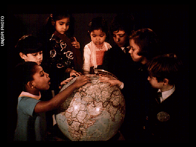 Seven children of diverse ethnicities stand circled around a globe as they point at different countries