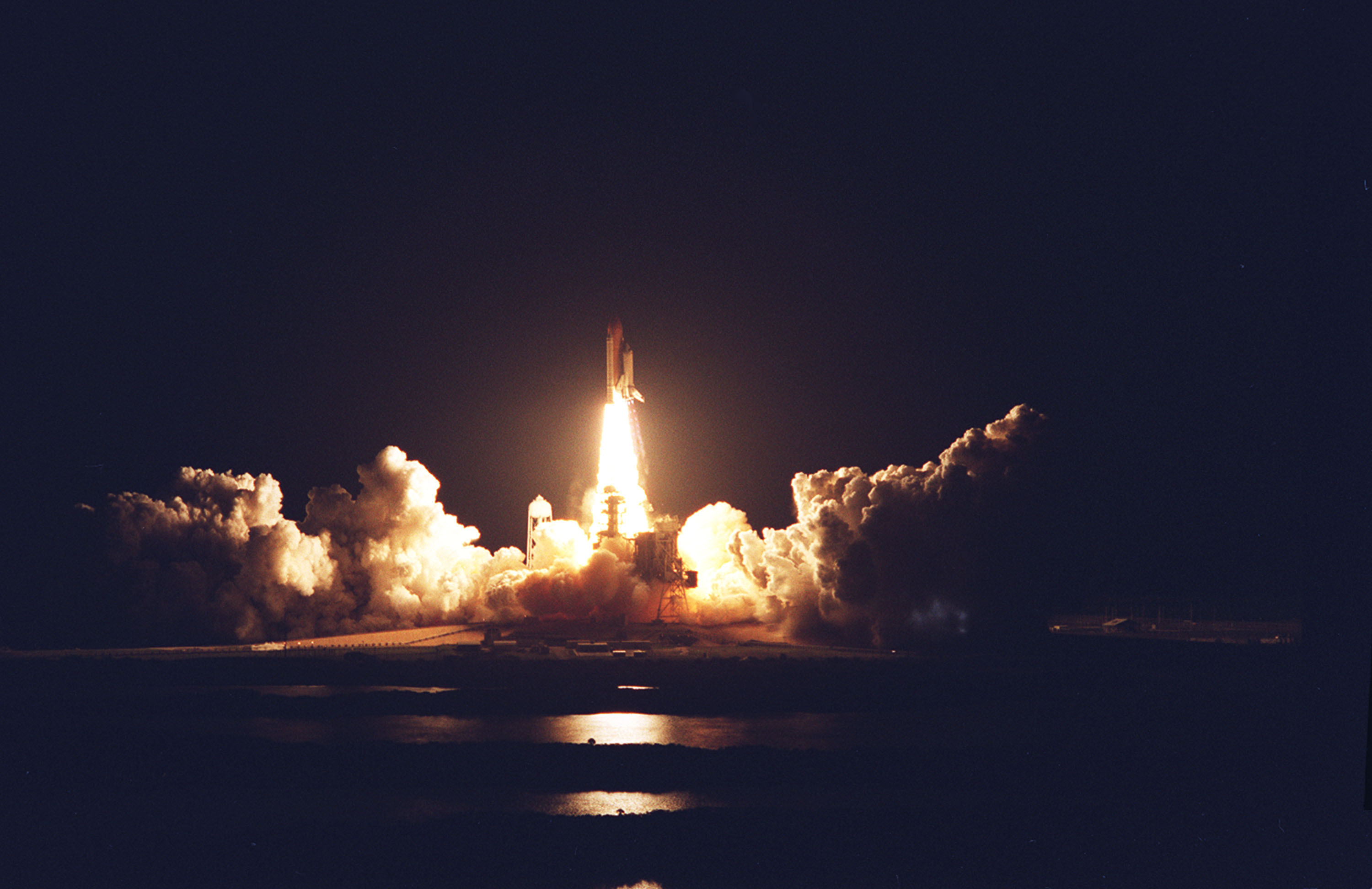 The space shuttle Columbia lifts off into a dark sky, illuminated by the flame pouring from its engine, which reflects as well off the clouds of billowing smoke surrounding it.