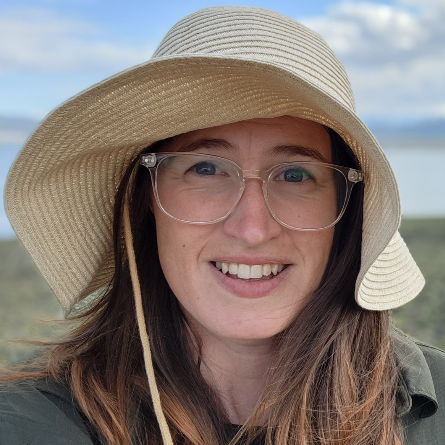 Close-up of a smiling person, wearing a sun hat and eyeglasses, outdoors under a partly cloudy sky.