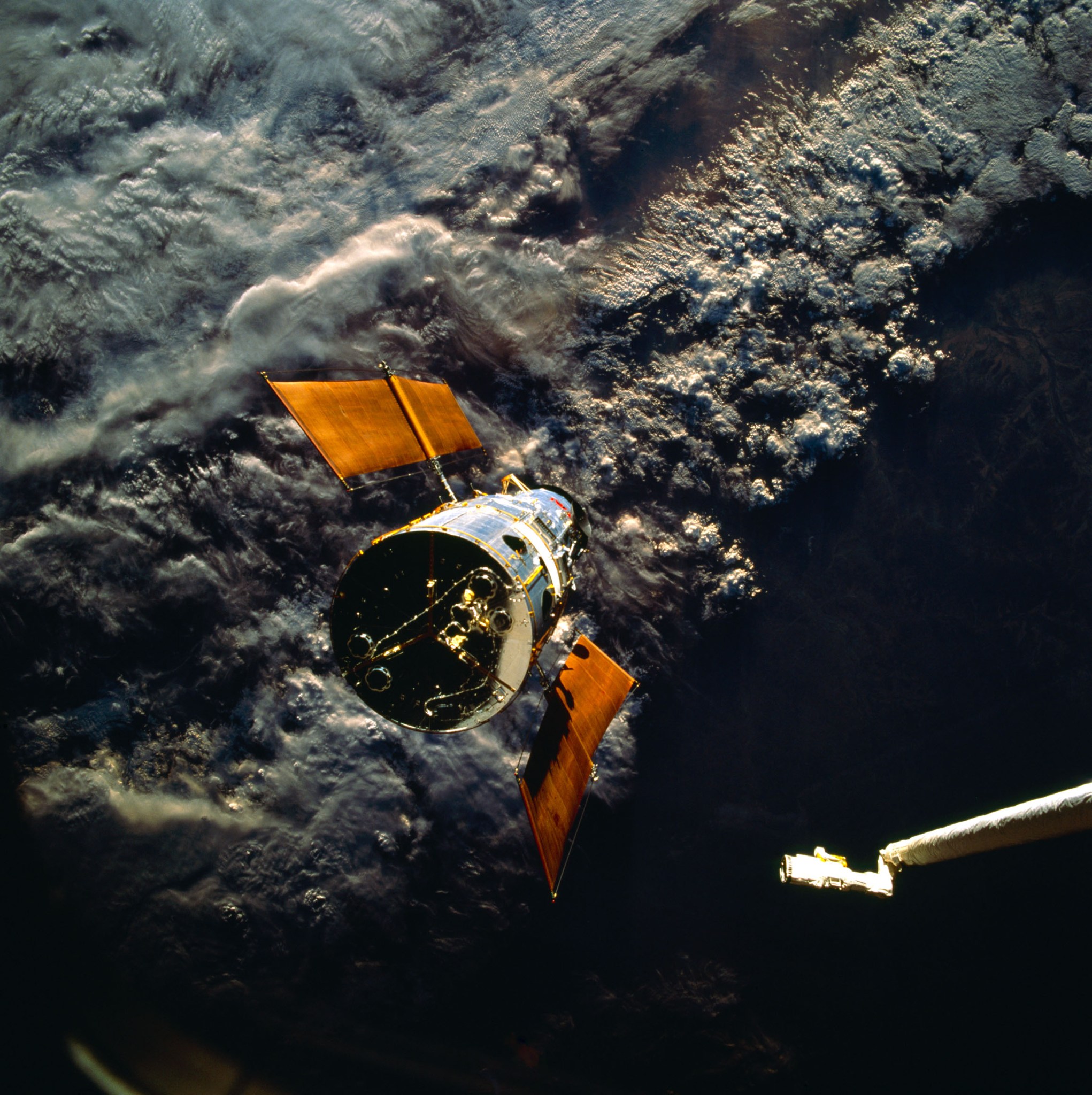 Hubble floats over a dark and cloudy Australia, oriented so the back of the telescope faces the viewer. A portion of the shuttle's robotic arm can be seen in the right corner, extending toward the telescope.