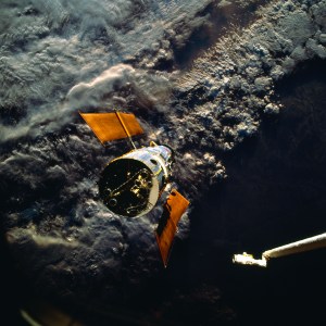 Hubble floats over a dark and cloudy Australia, oriented so the back of the telescope faces the viewer. A portion of the shuttle's robotic arm can be seen in the right corner, extending toward the telescope.