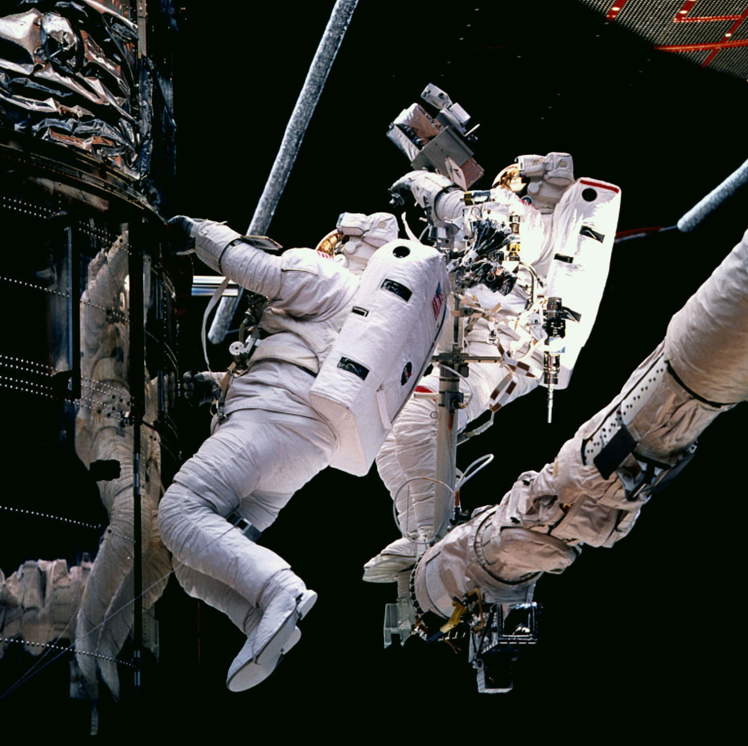 Two astronauts work on the Hubble telescope. One grips onto the telescope and the other is standing on the robotic arm.