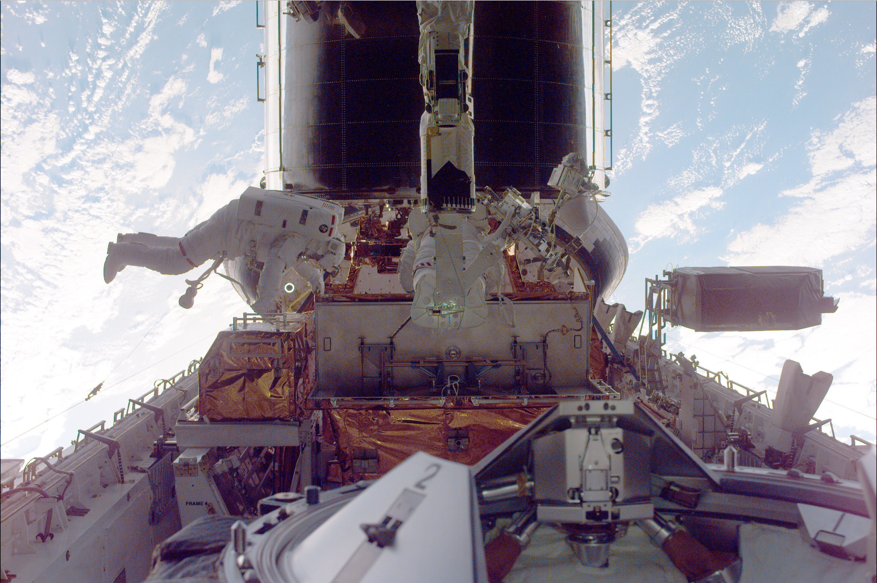 Earth fills the background behind Hubble in its position in the space shuttle's cargo bay. Two astronauts work in the cargo bay, one partially obscured by the shuttle's robotic arm.