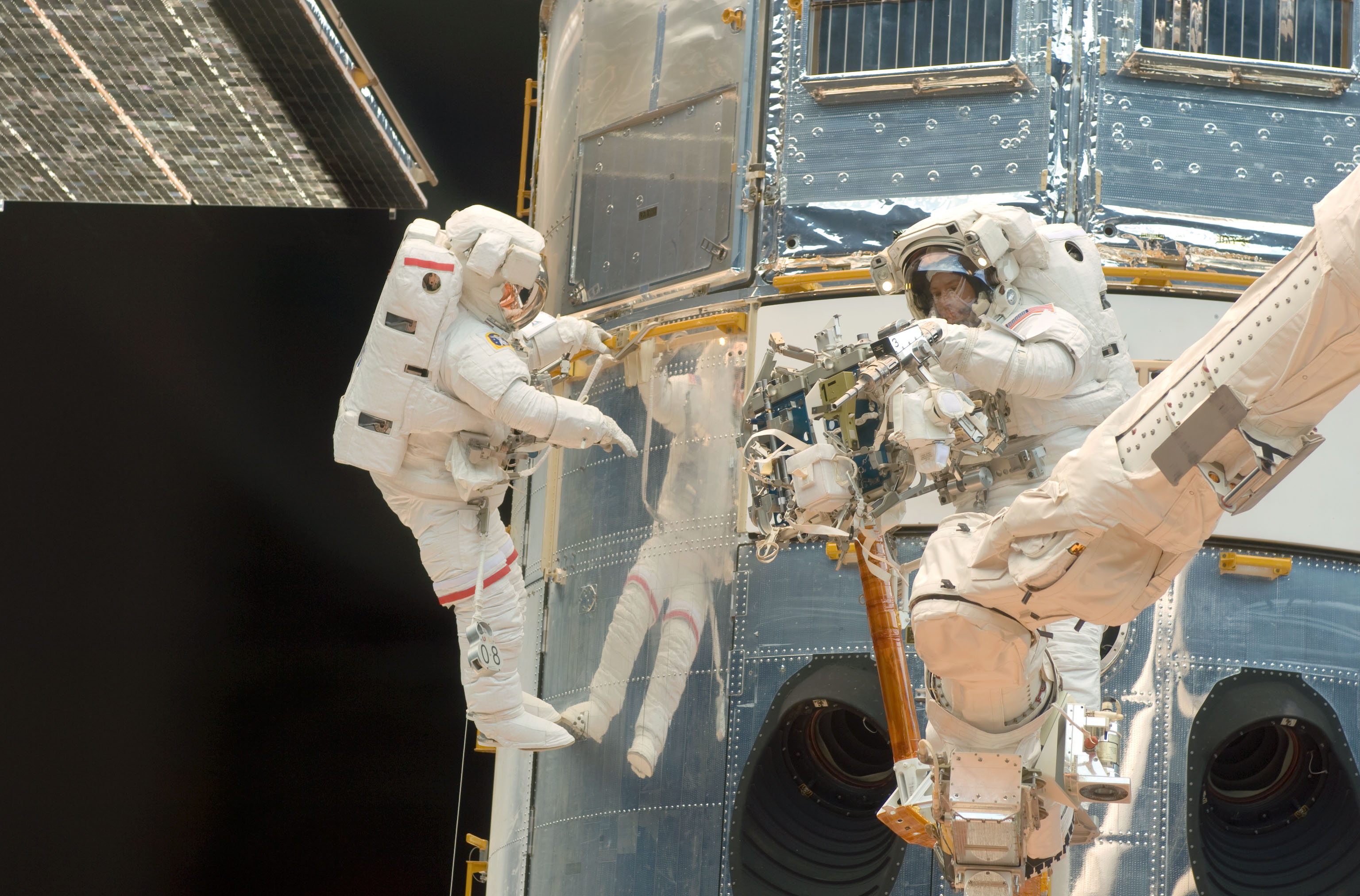 One astronaut holds to a yellow handrail on the Hubble telescope while another, perched on the shuttle's robotic arm, examines a drill-like power tool.