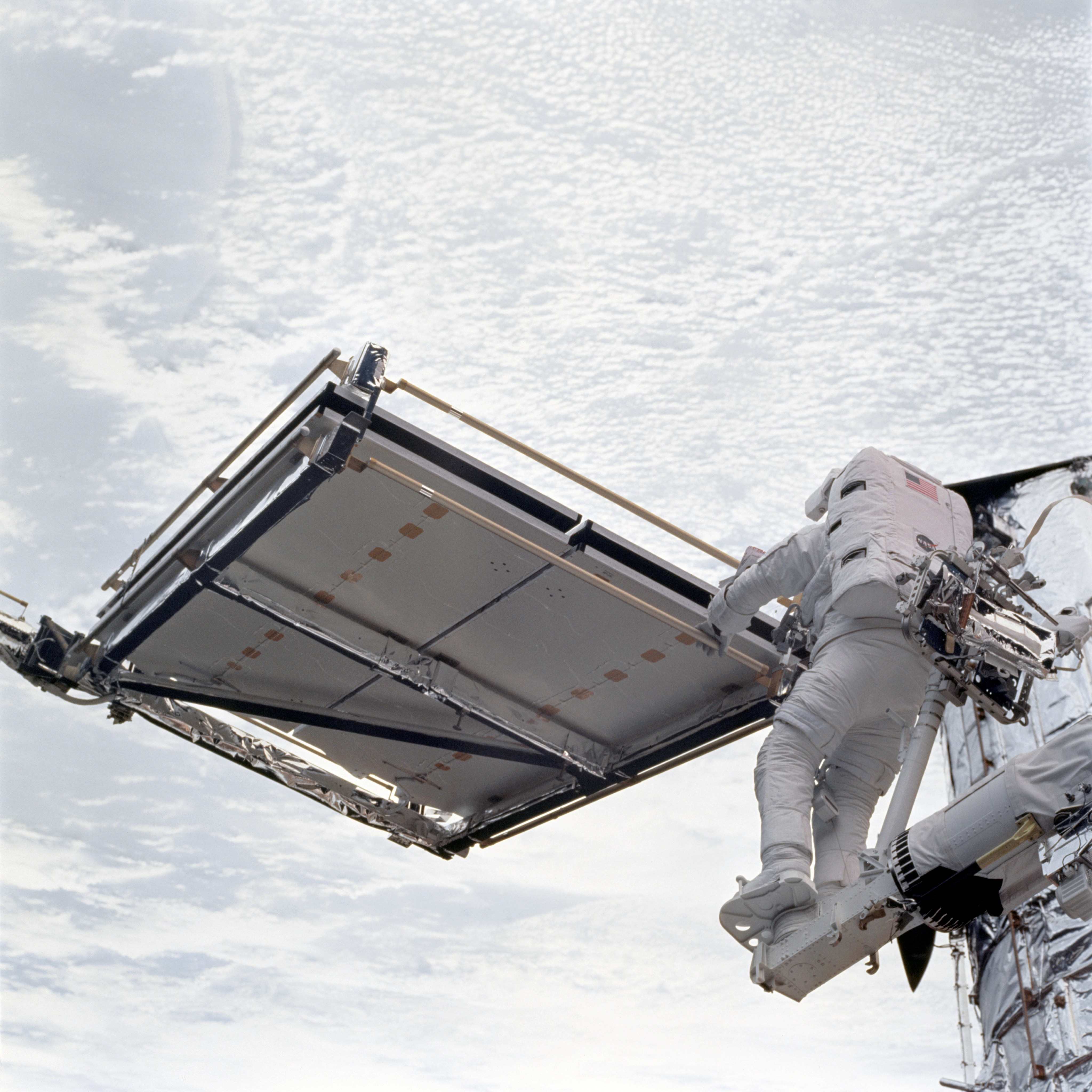 An astronaut works on a flat, square-shaped device (a folded solar array) against the white cloudy background of Earth.