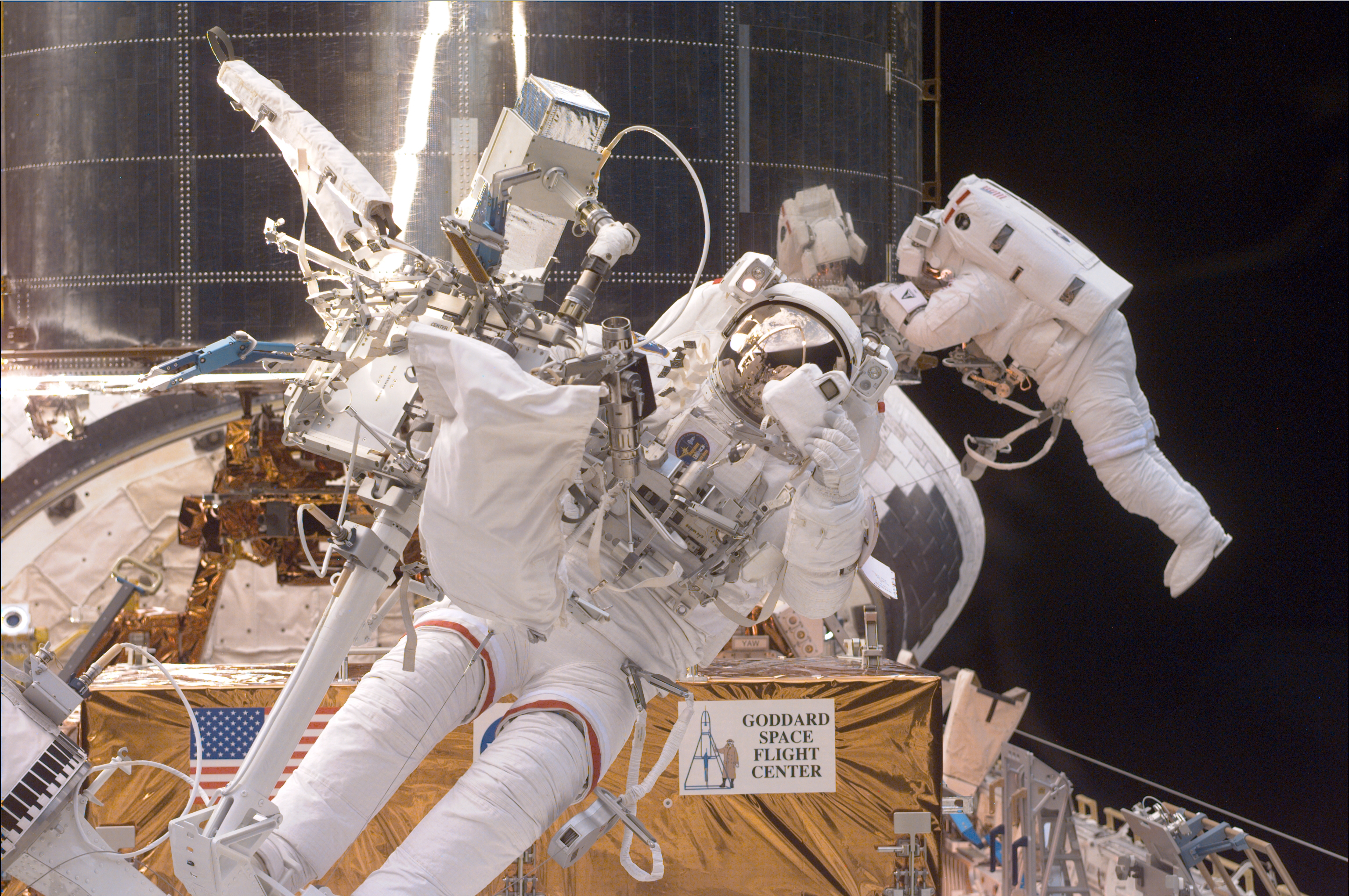 A astronaut anchored to the robotic arm in front of Hubble holds up a camera device to take a picture. Another astronaut is visible working on Hubble behind him.