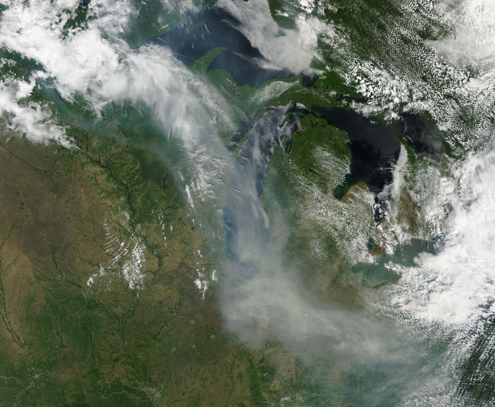 A satellite view shows the greens and orange browns of land in the Midwest of the United States. The land, split up by dark blue lakes at the top of the image, also has thin twisting rivers snaking through it. The land and water take up the background of the image. In the foreground are wisps of white clouds and gray smoke. Clouds are seen in the top left corner of the image and alone the right side of the image. The smoke trails from the top left of the image to the bottom right, and the land can be seen through the smoky plumes.