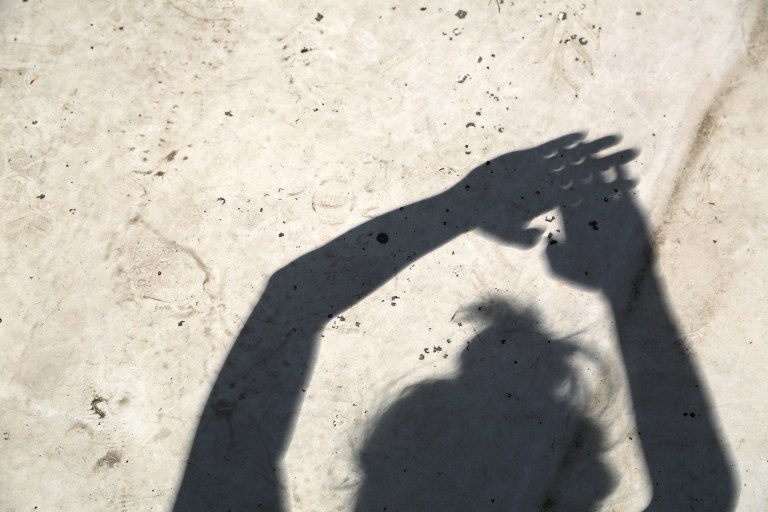 The shadow of a woman is seen on white concrete. She is holding her hands up, with her fingers crossed. Between the gaps in her fingers are the crescents of a partial solar eclipse