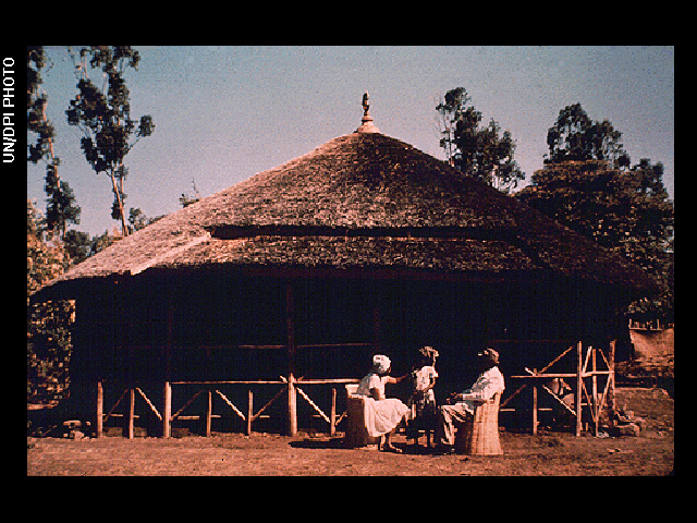 A small house with a grass roof and a fence made of sticks, located in a desert area with three people sitting near the front door