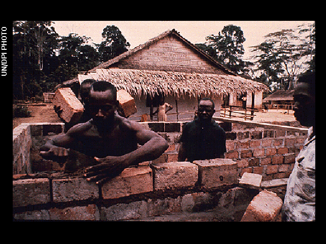 Four men work to lay bricks to build a small structure next to a second building that has a grass roof