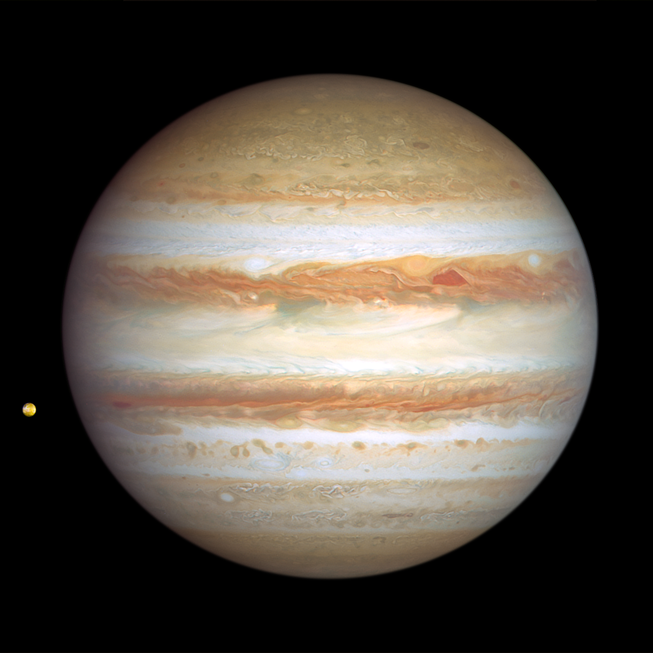Image of Jupiter taken on January 6, 2024. This side of Jupiter is also banded in stripes of brownish orange, light gray, soft yellow, and shades of cream, with many large storms and small white clouds punctuating the planet. At upper right of center, a pair of storms appear next to each other: a deep-red, triangle-shaped cyclone and a reddish anticyclone. Toward the far-left edge of this view is Jupiter's tiny orange-colored moon Io.