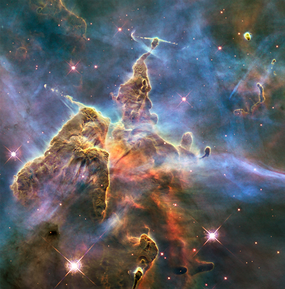 The Mystic Mountain is seen as a chaotic pillar of colorful gas and dust, narrowing toward the top of the image. The dust and gas is mostly yellow, brown, and orange, all jutting against a hazy purple and blue background with a few pink stars.