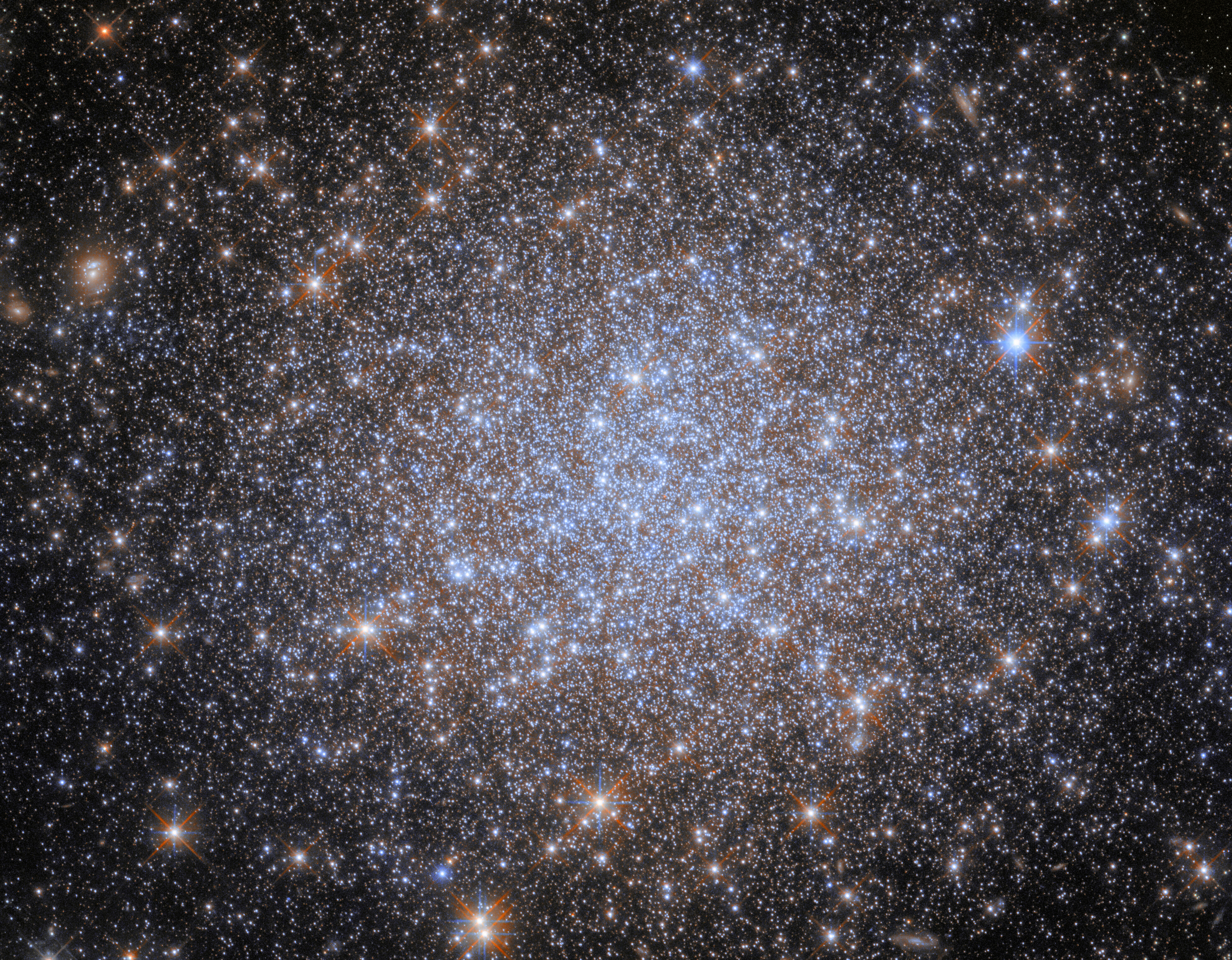 A cluster of stars. Most of the stars are very small and uniform in size, and they are notably bluish and cluster more densely together toward the center of the image. Some appear larger in the foreground. The stars give way to a dark background at the corners.