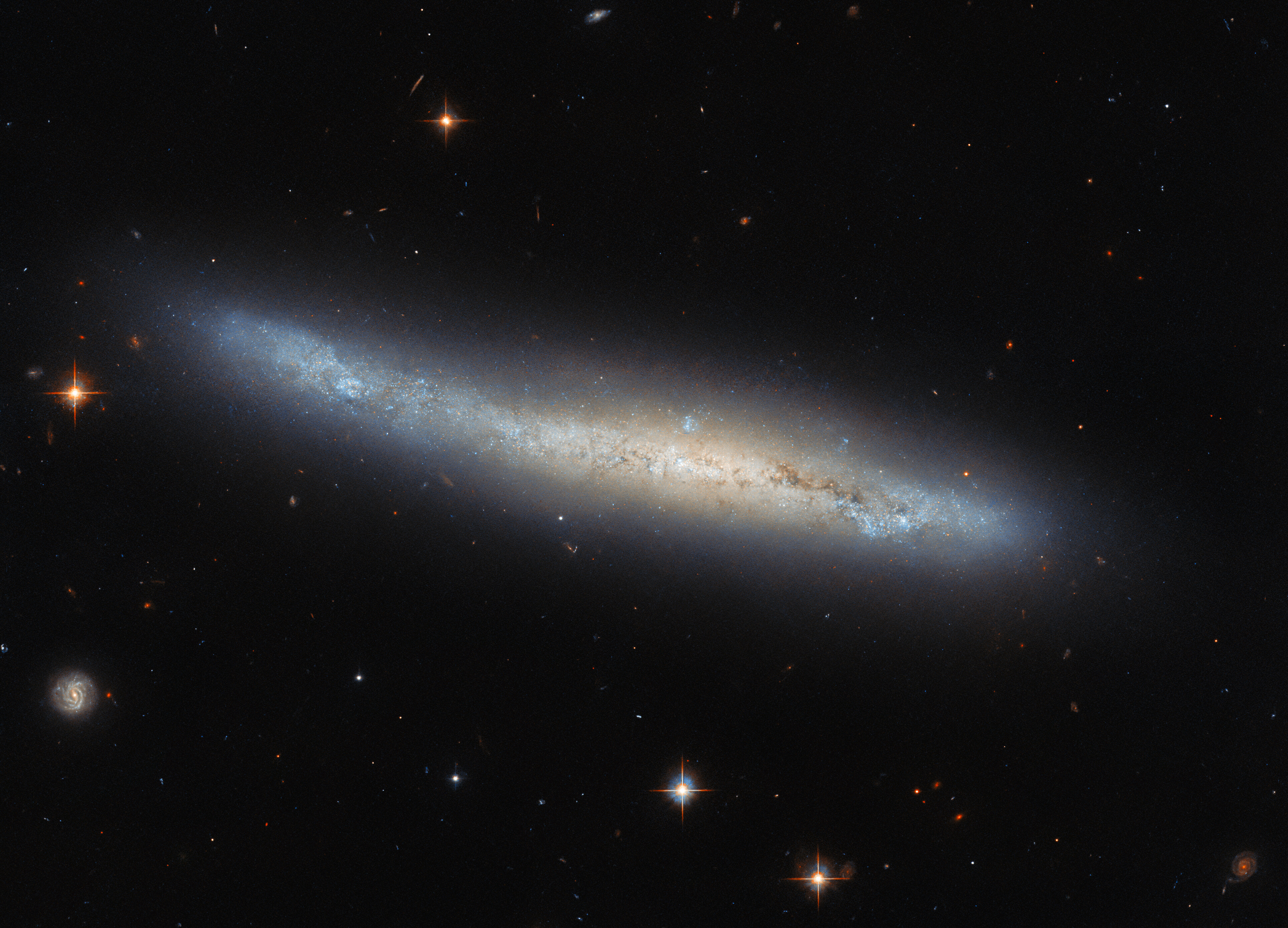 A broad spiral galaxy is seen edge-on, so that its spiral arms can’t be seen. Visible dust and stars trace the disk of the galaxy, surrounded by a glowing halo above and below. The color of the galaxy changes smoothly between the outer disk at the ends and the bulge in the center. A few bright stars surround the galaxy on a dark background.