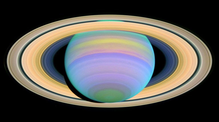 Saturn, seen in purples, greens and blues, is surrounded by its bright yellow rings.