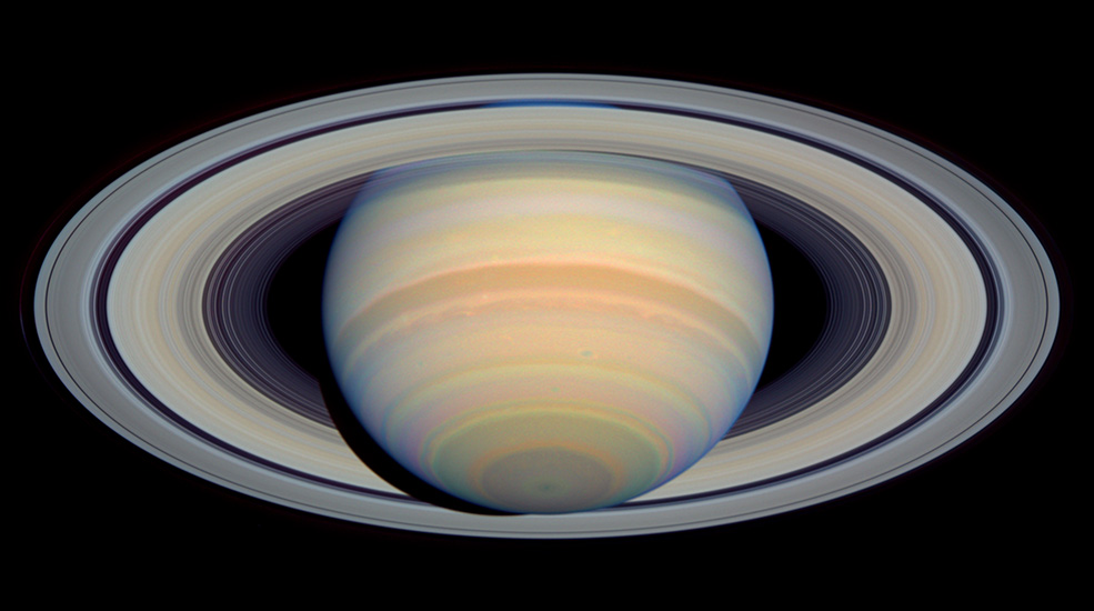 Saturn and its rings. The planet appears as though it is tilted backward, appearing to reveal the underside of its rings. Overall Saturn is yellow with bands of red, yellowish-brown, light orange, pink, and blue.