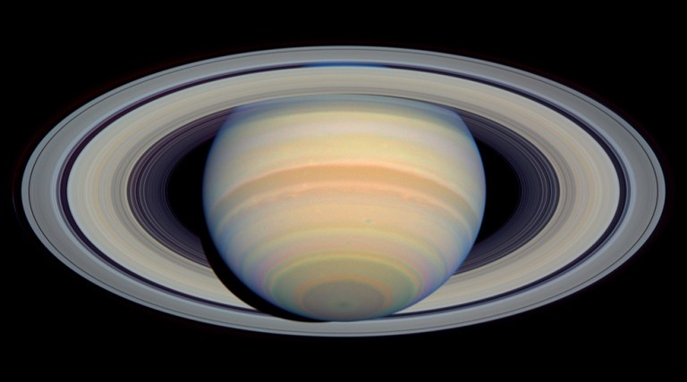 Saturn and its rings. The planet appears as though it is tilted backward, appearing to reveal the underside of its rings. Overall Saturn is yellow with bands of red, yellowish-brown, light orange, pink, and blue.