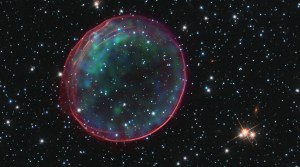 Black background dotted with stars. A dark-red transparent bubble of gas with blue and green looking like a ring. Stars are visible through the center of the ring.