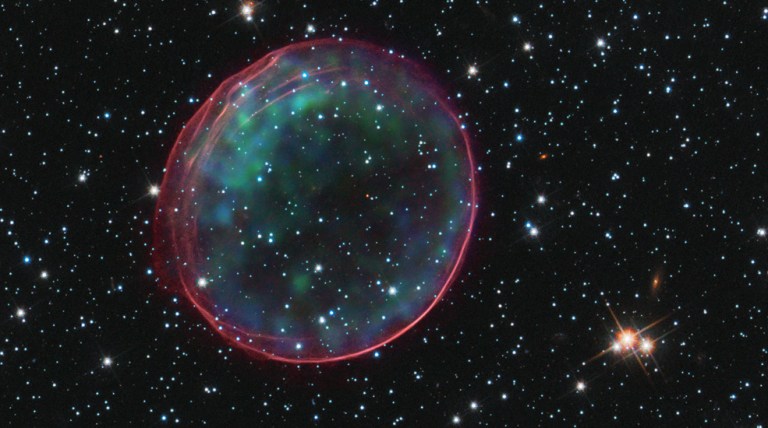 Black background dotted with stars. A dark-red transparent bubble of gas with blue and green looking like a ring. Stars are visible through the center of the ring.