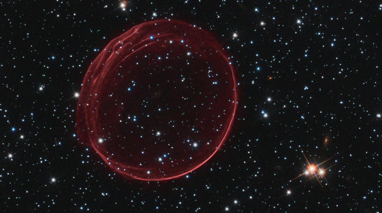 Black background dotted with stars. A dark-red transparent bubble of gas looking like a ring. Stars are visible through the center of the ring.