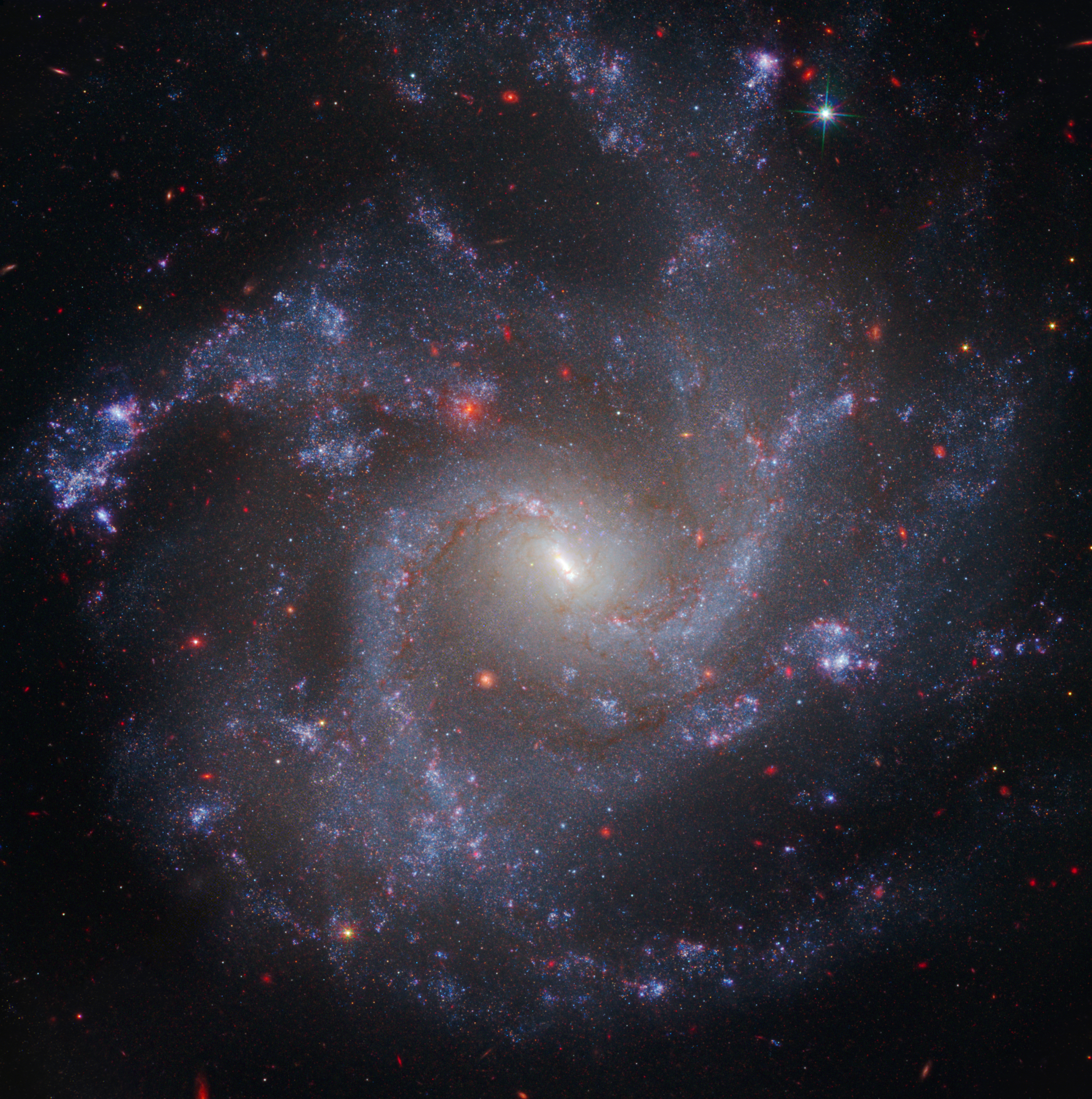 A spiral galaxy with a small bar of bright-white stars at its core. Two main spiral arms extend outward from each end of the bar. They appear to fork into multiple branches beyond the galaxy's core. The spiral arms have a lavender hue. Bright-white and bright-red stars dot the galaxy. Reddish-brown dust lanes line the inner curves of the spiral arms.