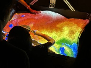 A girl uses a shovel to move sand and change a terrain visualization illuminated by colors representing elevation.