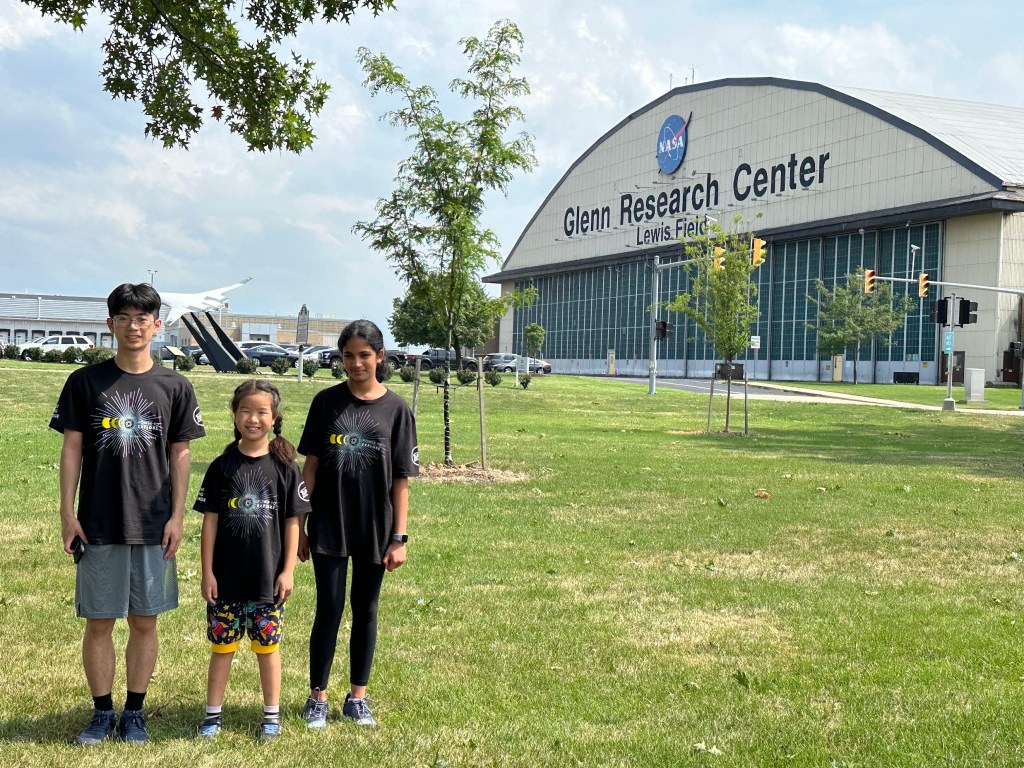 Three students stand in a grassy field in front of a hangar that says NASA Glenn Research Center.