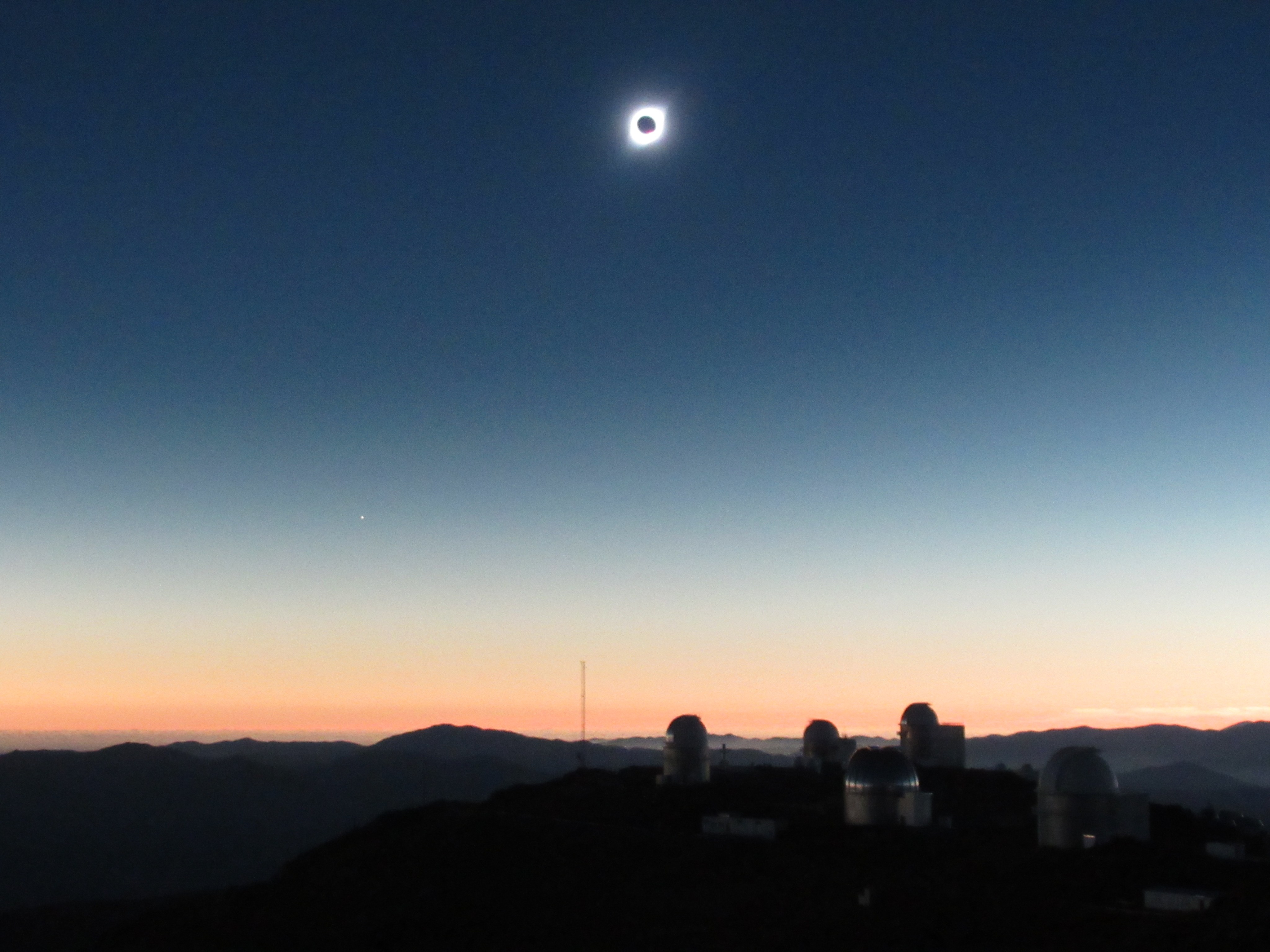A total solar eclipse is seen high in the sky against a sky that fades from dark blue, to light blue, to light orange and pink. Closer to the landscape a faint white dot is there – Venus.