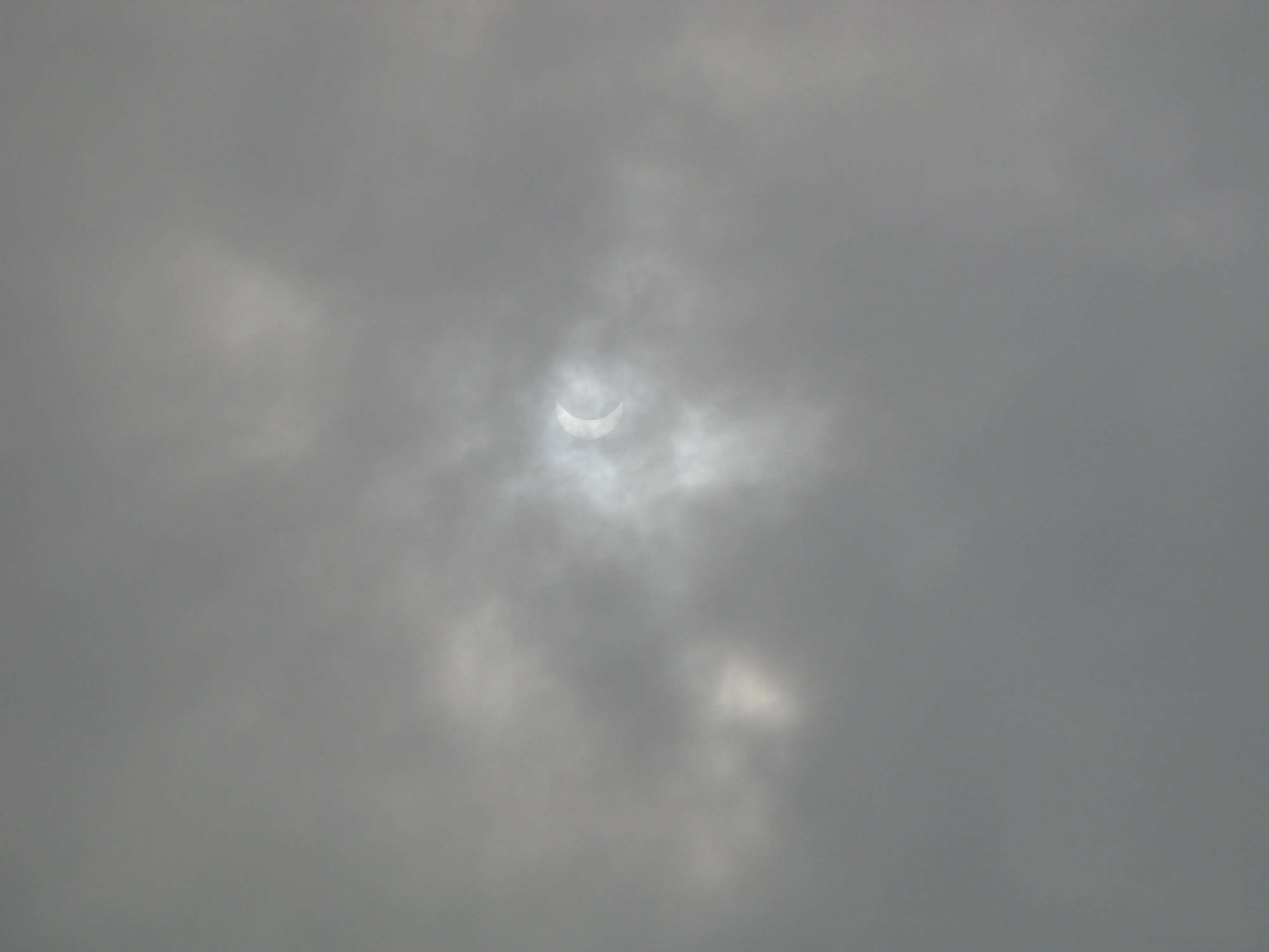 The partially eclipsed Sun can barely be seen as a white crescent through clouds.