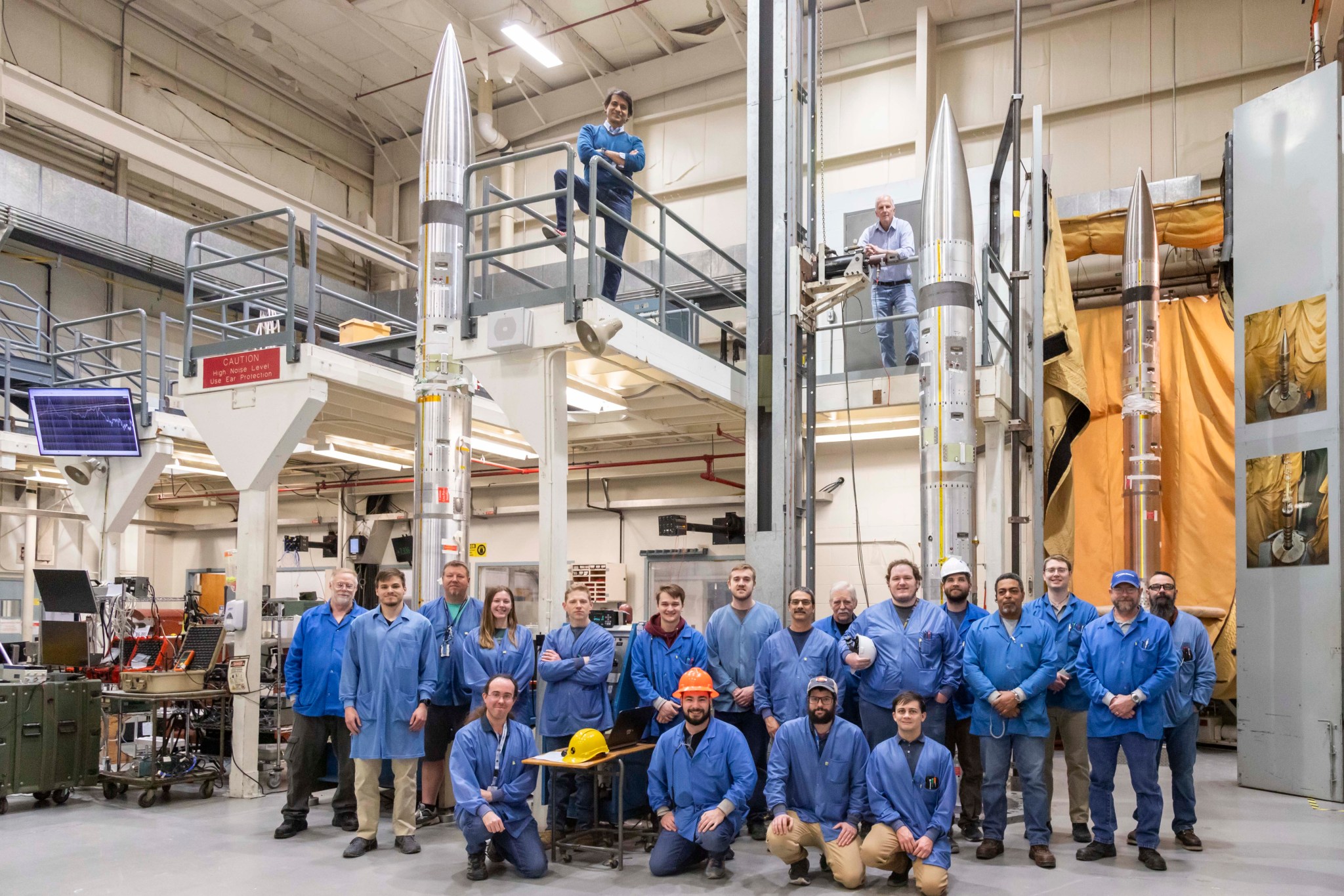 A group of people wearing blue jackets pose for the picture. They stand inside a tall, industrial room. Three silver rockets are behind them.