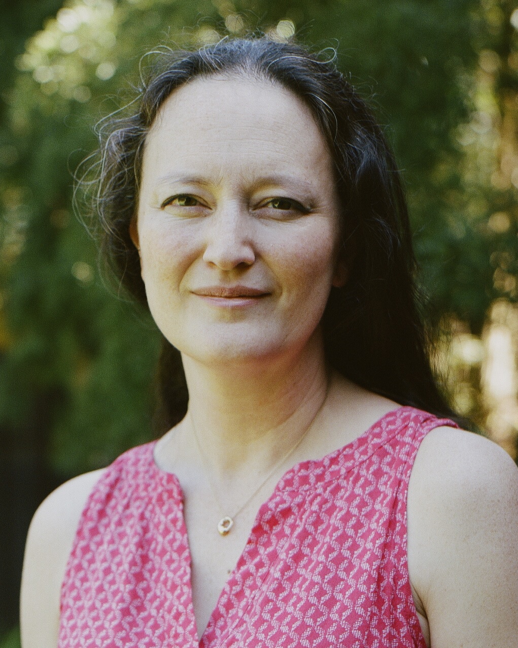 Portrait photo of a smiling dark haired woman outdoors with trees in the background.