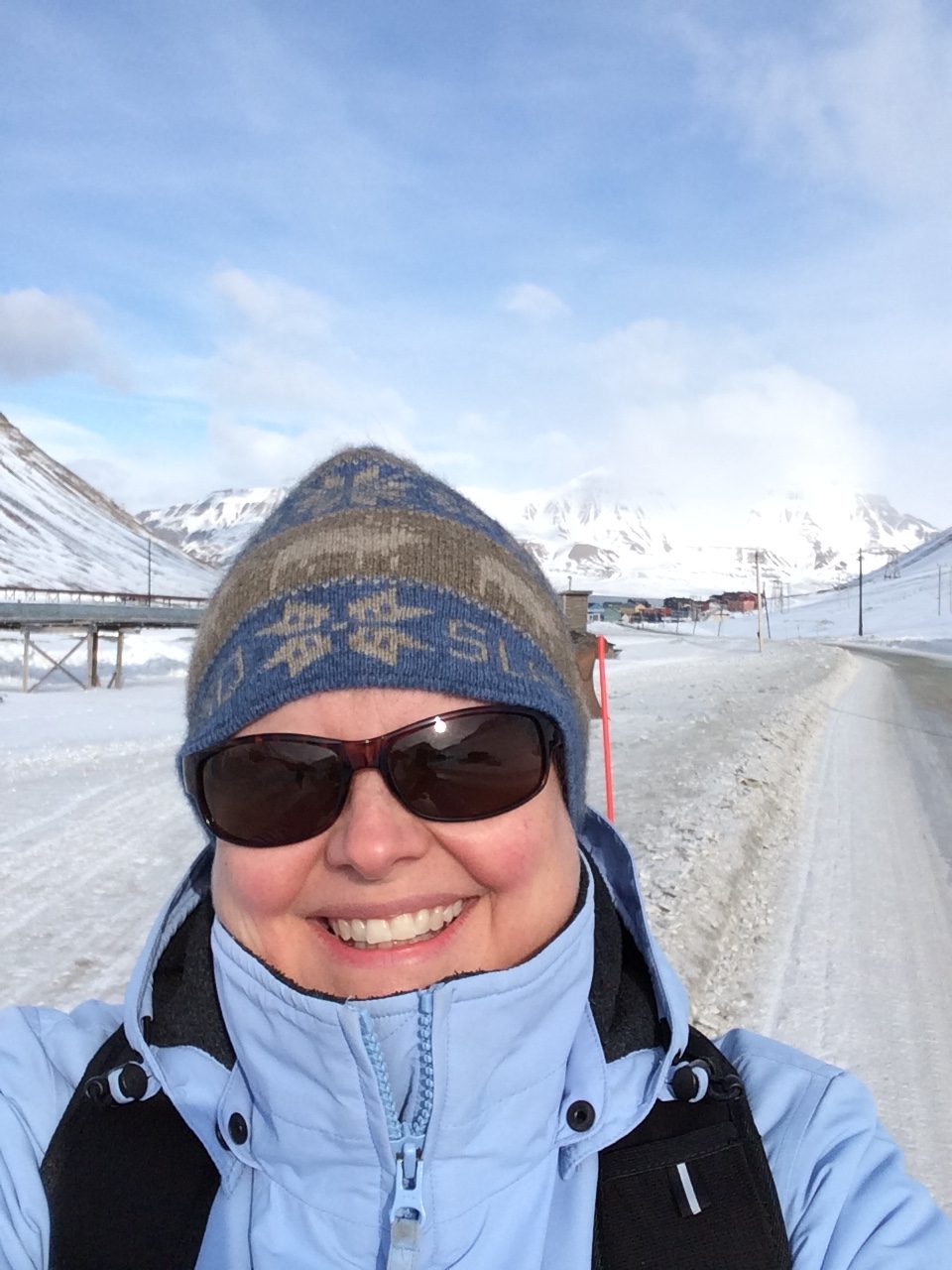 A selfie of a woman in a blue coat, knitted beanie, and sunglasses standing in front of an icy background. She appears to be standing on the side of a slushy, cleared road and snow-covered mountains and cloudy blue skies are behind her.