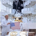 A man holds an American Flag over the Voyager Golden Record in a cleanroom. The completed Voyager Spacecraft is in the background.