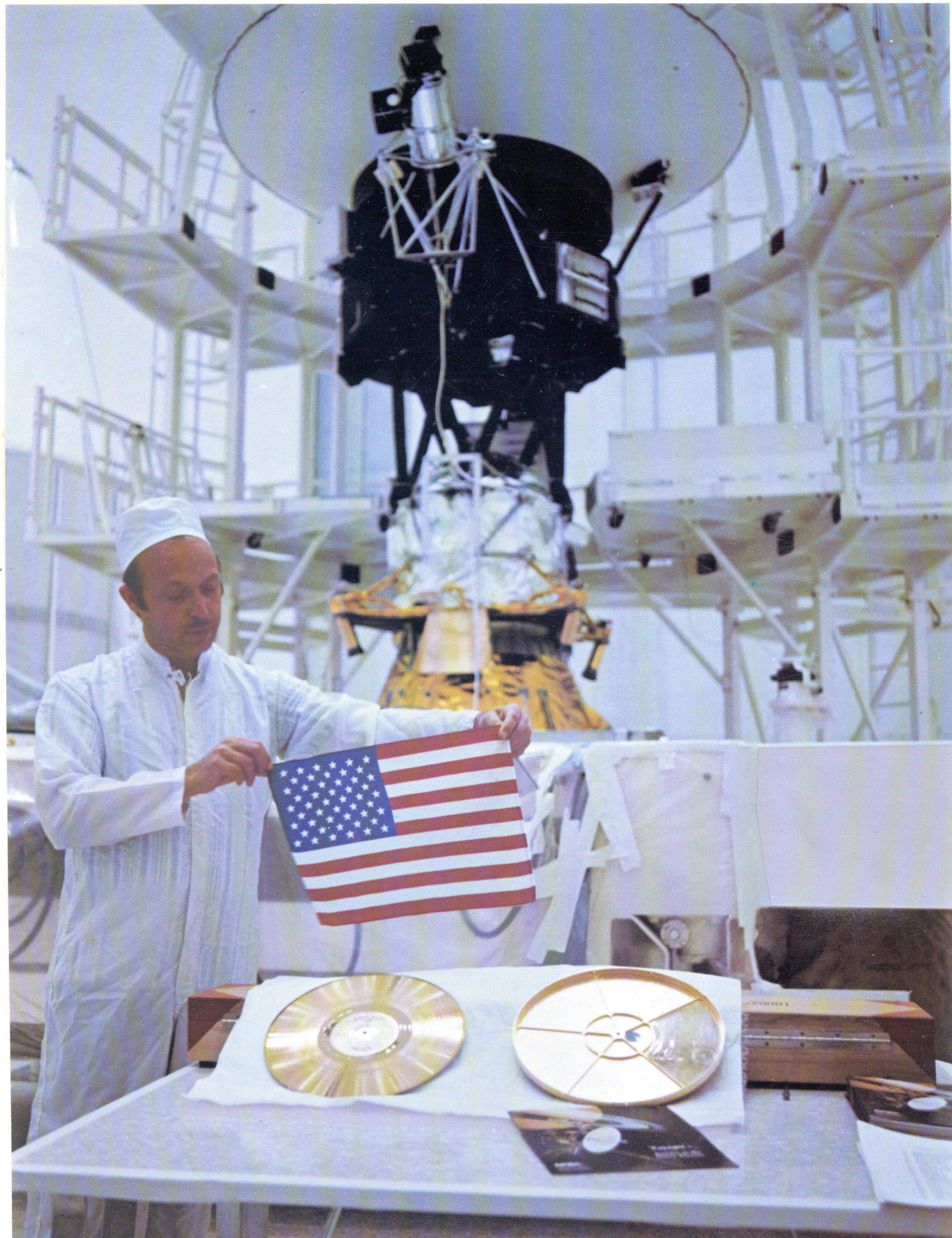 A man holds an American Flag over the Voyager Golden Record in a cleanroom. The completed Voyager Spacecraft is in the background.